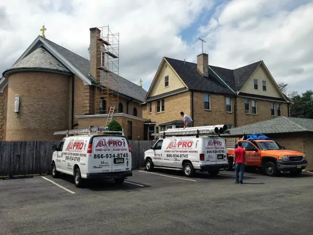 Two ALL PRO work vans and a pickup parked at a job site beside a large brick building