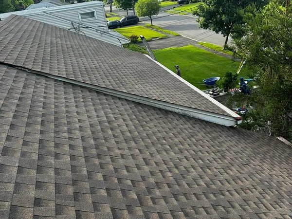 Completed roof with weathered-brown architectural shingles above a tree-lined street