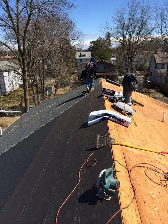 Two roofers shingling one side of a roof while the other side shows new plywood decking