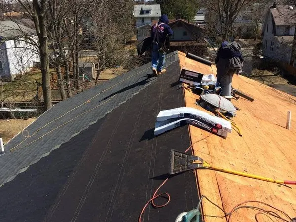 Two roofers shingling one side of a roof while the other side shows new plywood decking