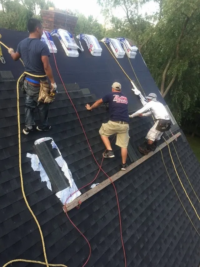 Three roofers working on a steep roof with safety ropes, with shingle bundles lined up at the ridge