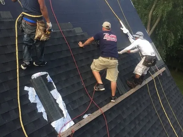 Three roofers working on a steep roof with safety ropes, with shingle bundles lined up at the ridge