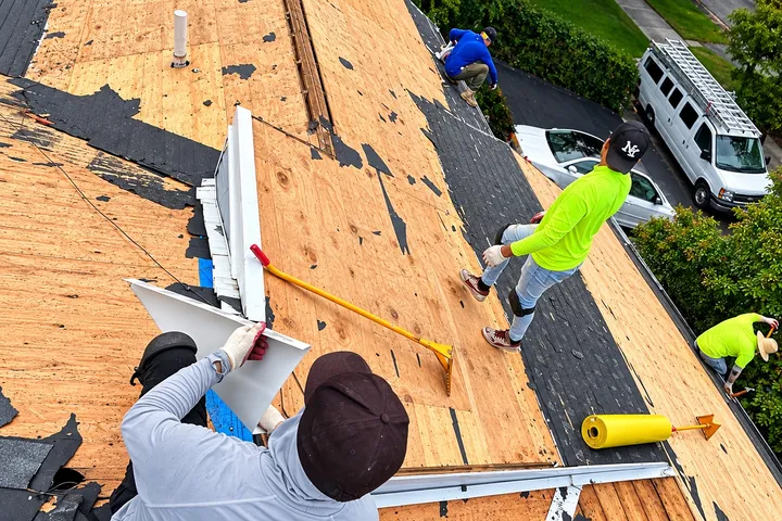 Four roofers tearing off old shingles and drip edge down to the plywood deck, with a work van parked below