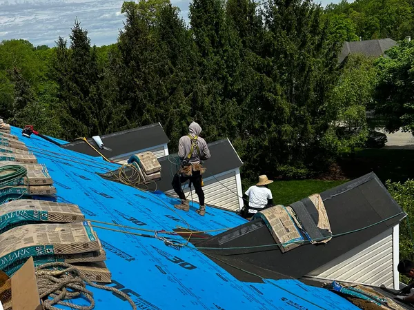 Two roofers working on a roof covered in blue synthetic underlayment, with shingle bundles staged at the edge