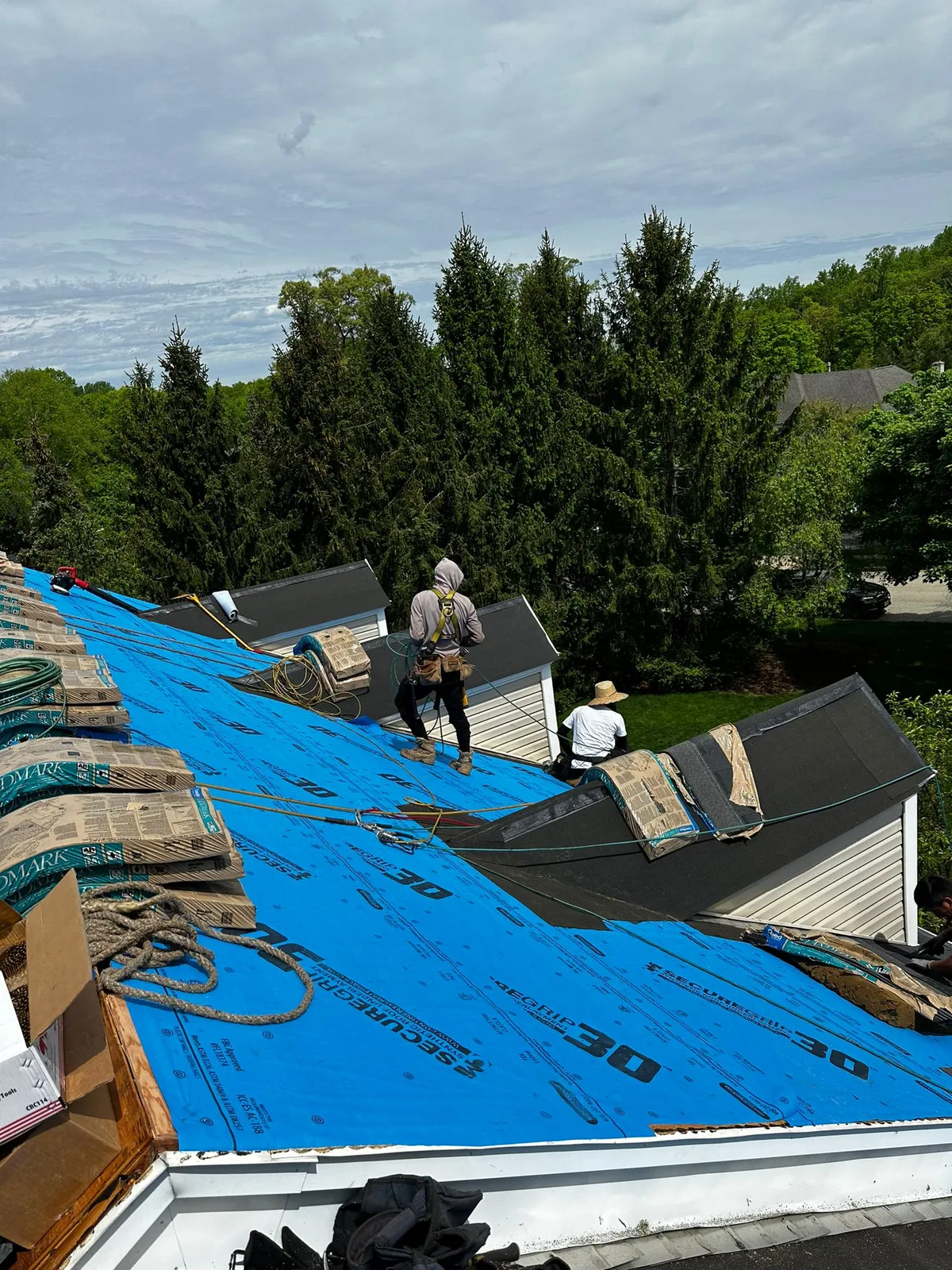Two roofers working on a roof covered in blue synthetic underlayment, with shingle bundles staged at the edge