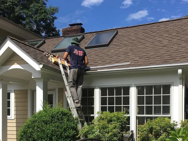 Roofer on a ladder working at the eave of a brown shingle roof with skylights