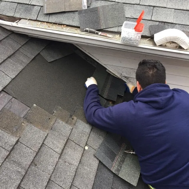 Roofer replacing damaged shingles at the roof edge above a gutter