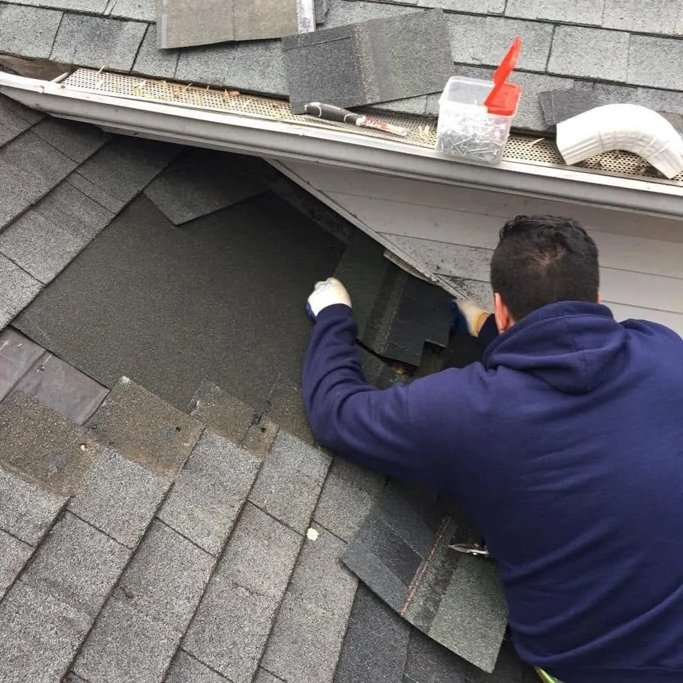 Roofer replacing damaged shingles at the roof edge above a gutter