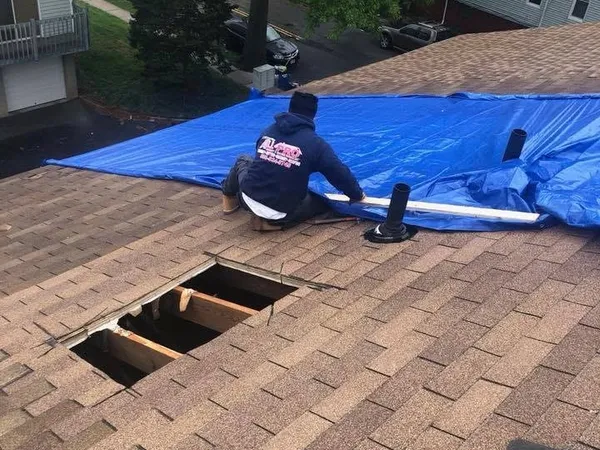 Roofer securing a blue tarp over a damaged section of roof, with open holes in the decking beside him