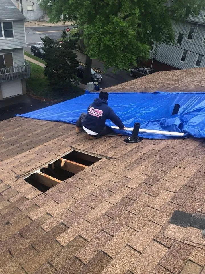 Roofer securing a blue tarp over a damaged section of roof, with open holes in the decking beside him