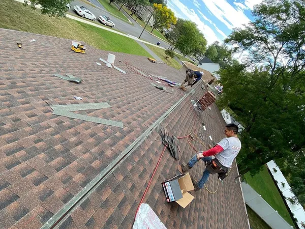 Two roofers installing new brown architectural shingles on a long roof slope
