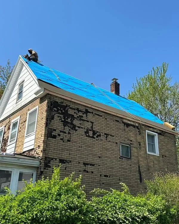 Blue tarp secured over a house roof for temporary protection, with a roofer working at the peak