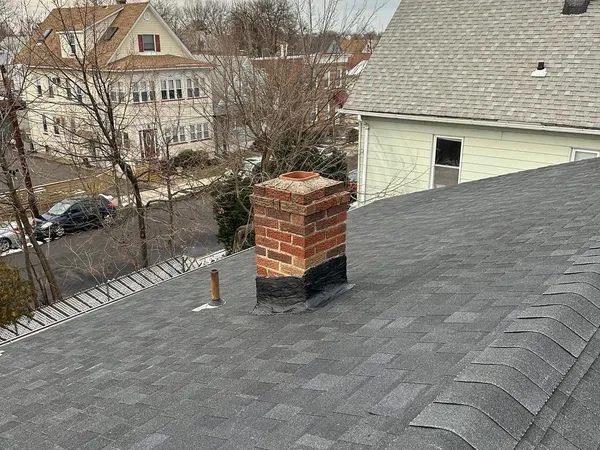 New dark gray shingle roof with a brick chimney and vent pipe, above a street of older homes