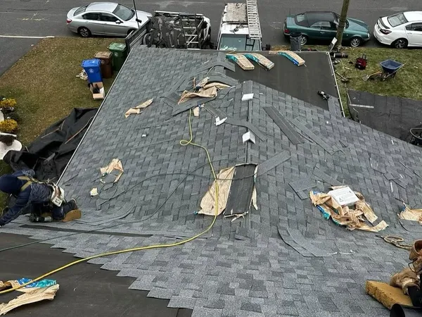 Gray shingle roof with torn and loose shingle pieces scattered across it, crew working at the edge