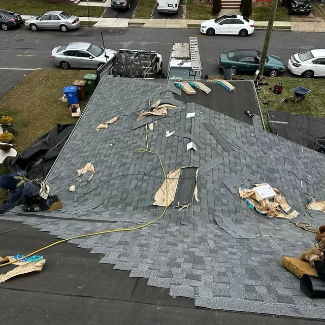 Gray shingle roof with torn and loose shingle pieces scattered across it, crew working at the edge