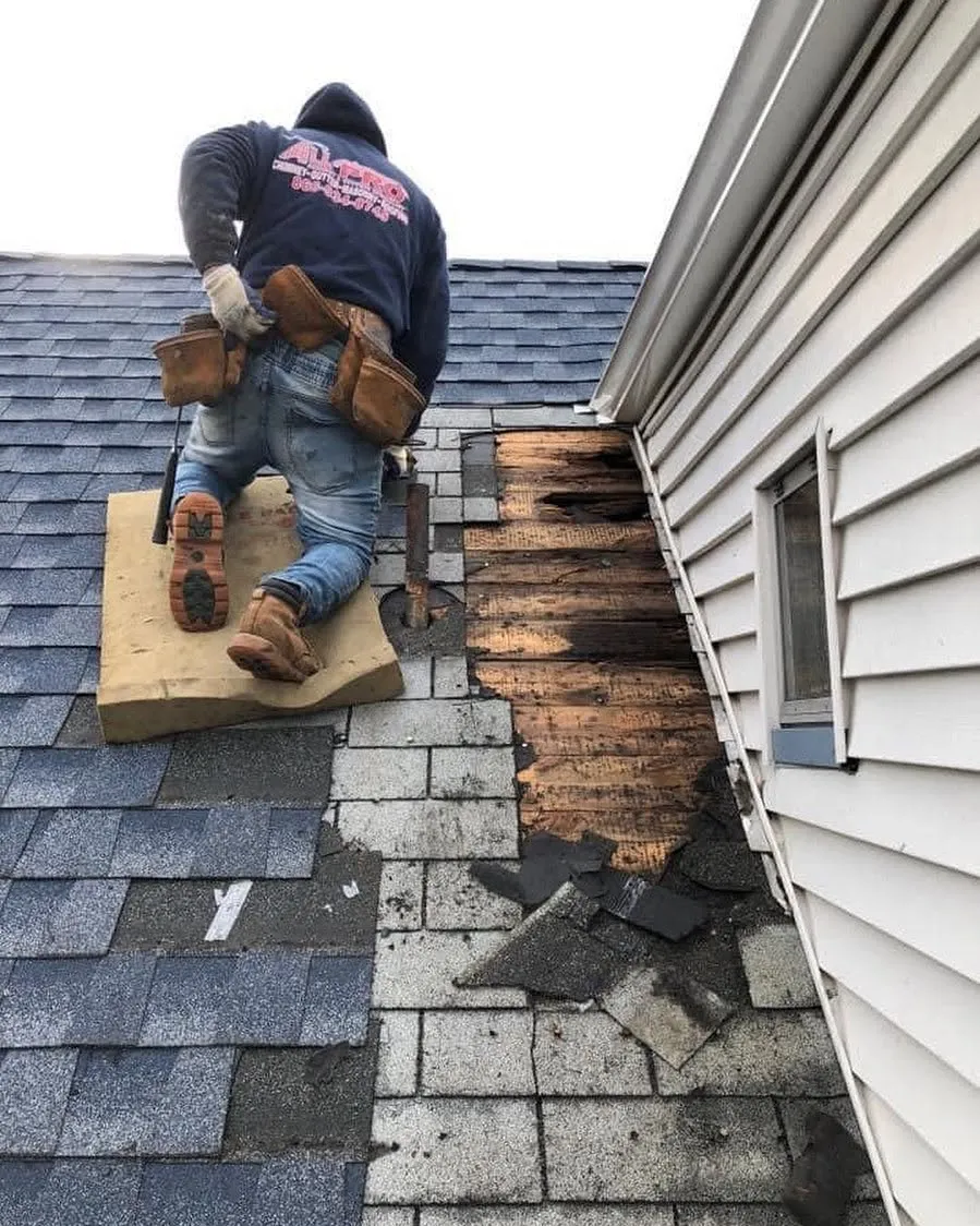 Roofer kneeling on a gray shingle roof beside a strip of rotted decking exposed along a wall