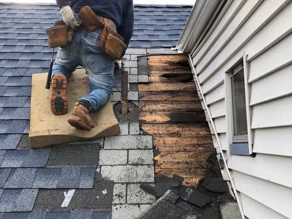 Roofer kneeling on a gray shingle roof beside a strip of rotted decking exposed along a wall