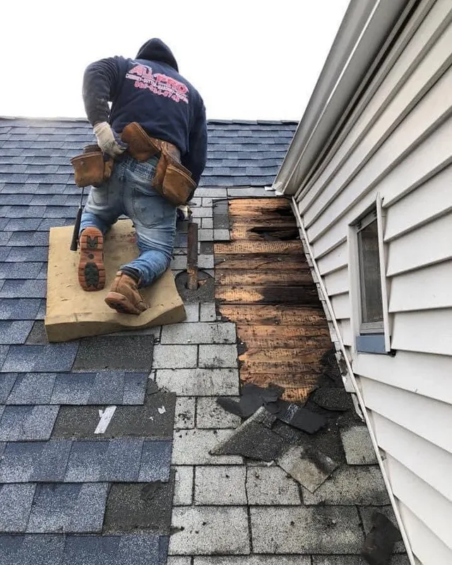 Roofer kneeling on a gray shingle roof beside a strip of rotted decking exposed along a wall