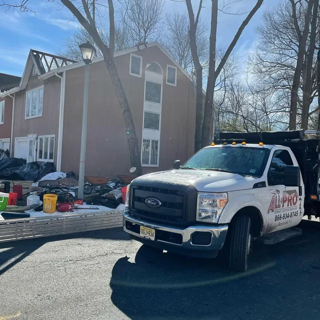 White ALL PRO dump truck parked at a job site beside a two-story home, with tear-off debris in the driveway
